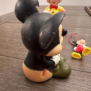 May include: Three vintage Mickey Mouse figurines. The largest figurine is black and green, sitting down with its hands clasped. The middle figurine is black and red, standing with its arms at its sides. The smallest figurine is black and yellow, lying on its side with its arms outstretched.