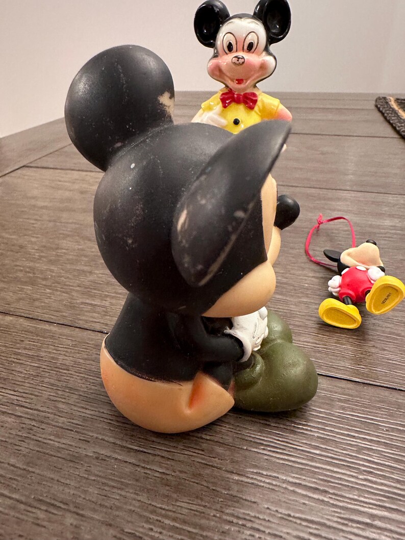 May include: Three vintage Mickey Mouse figurines. The largest figurine is black and green, sitting down with its hands clasped. The middle figurine is black and red, standing with its arms at its sides. The smallest figurine is black and yellow, lying on its side with its arms outstretched.