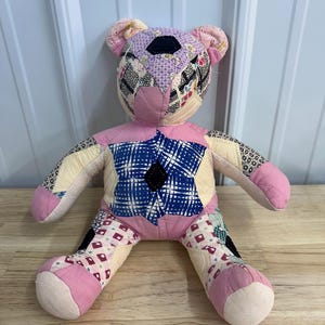 May include: A handmade patchwork teddy bear with a pink body, limbs, and ears. The head features a patchwork design with various fabric patterns and colors, including pink, purple, and black. The bear's chest has a blue and white patterned design with a black diamond.