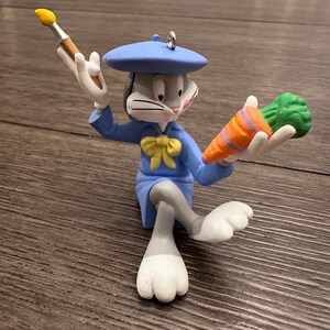 May include: A gray rabbit wearing a blue beret and a blue dress with a yellow bow holds a paintbrush in one hand and a carrot in the other.