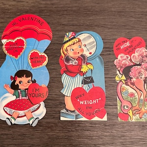 May include: Three vintage Valentine's Day cards with red hearts and messages. The first card features a red parachute with a heart that says "Just Dropping In With My Heart". The second card features a girl in a red dress and a heart that says "I'm Yours". The third card features a girl in a red dress and a heart that says "Why "Weight" I'm All Yours".