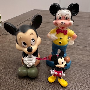 May include: Three vintage Mickey Mouse figurines. The largest figurine is standing with a yellow shirt, blue trousers, and a red bow tie. The middle figurine is sitting with its hands clasped. The smallest figurine is standing with red shorts and yellow shoes.