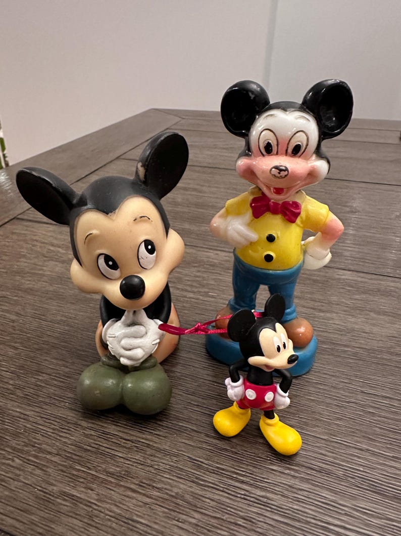 May include: Three vintage Mickey Mouse figurines. The largest figurine is standing with a yellow shirt, blue trousers, and a red bow tie. The middle figurine is sitting with its hands clasped. The smallest figurine is standing with red shorts and yellow shoes.