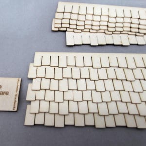May include: Wooden miniature roof shingles and a small square piece with the text "One Square Inch". The shingles are light-colored and arranged in rows, suggesting a roofing material for a model or craft project.