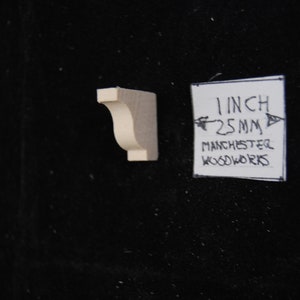 May include: A small, unfinished, wooden corbel with a 1-inch (25 mm) measurement. The corbel is a decorative architectural element used to support a shelf or other structure.