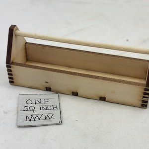Peut inclure: Petite boîte à outils en bois non finie avec une poignée. La boîte est rectangulaire, de couleur bois clair avec des accents marron foncé. Une petite carte carrée avec le texte "ONE 5Q INCH" est à côté.