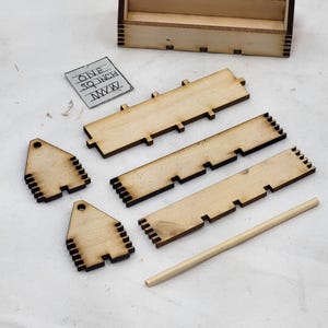 May include: A wooden box with a small wooden shelf inside. The box is disassembled and laid out on a white surface. The box is labeled "1 inch square" and includes a small wooden dowel.