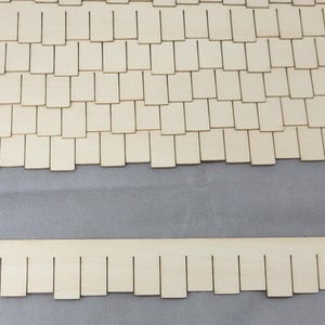 May include: Wooden miniature roof shingles and trim pieces. The shingles are rectangular and arranged in a staggered pattern. A small square piece is labeled "One Square Inch". The trim piece has a scalloped edge.