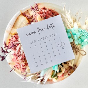 May include: A white card with the text "save the date" and a calendar for September 2026, with the 12th circled. The card is surrounded by dried flowers in shades of pink, cream, and teal, arranged on a wooden plate.