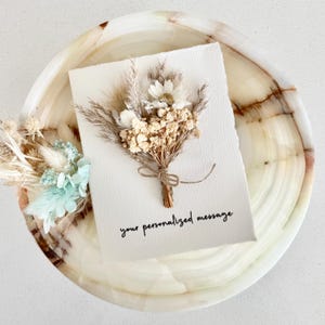 Puede incluir: Una tarjeta blanca con un ramo de flores secas atado con hilo. La tarjeta dice "your personalized message" en escritura negra. La tarjeta está sobre una bandeja de mármol.