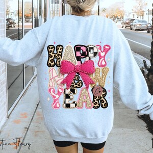 Pode incluir: Camisola cinza claro com as palavras "HAPPY NEW YEAR" em um design colorido. O design inclui estampa de leopardo, rosa, dourado e padr&otilde;es xadrez em preto e branco, com um grande la&ccedil;o rosa no centro.