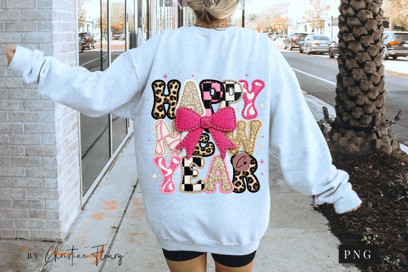 Pode incluir: Camisola cinza claro com as palavras "HAPPY NEW YEAR" em um design colorido. O design inclui estampa de leopardo, rosa, dourado e padr&otilde;es xadrez em preto e branco, com um grande la&ccedil;o rosa no centro.