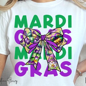 May include: White t-shirt with the words "Mardi Gras" in green and purple lettering. A purple bow with Mardi Gras themed decorations is in the centre of the shirt.