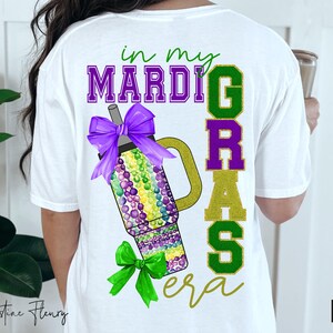 May include: White t-shirt with the text "in my Mardi Gras era" in green, purple, and gold glitter letters. A purple and green Mardi Gras cup with a bow is in the center of the shirt.