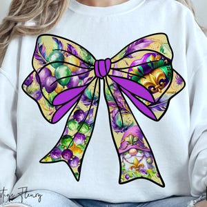 May include: A purple and gold bow with Mardi Gras beads, feathers, and a gnome design. The bow is on a white sweatshirt.