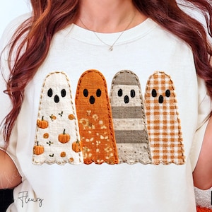 Può includere: T-shirt color crema con quattro figure di fantasmi applicate. Ogni fantasma ha un motivo diverso: zucche, floreale, righe e quadretti. I fantasmi hanno occhi e bocche neri. La maglietta è un design a tema autunnale.