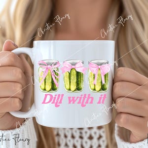 May include: White ceramic mug with a pink and green design of three jars of dill pickles with pink bows. The text "Dill with it" is printed in pink below the jars.