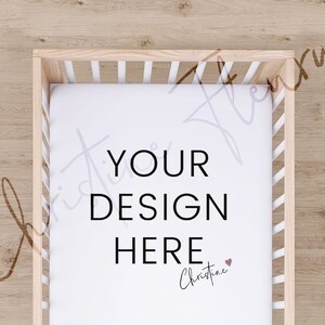 May include: A white cot sheet with the text "YOUR DESIGN HERE" and a small heart with the name "Christine" written below. The sheet is in a wooden cot with white slats.