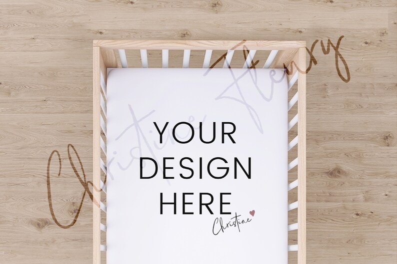 May include: A white cot sheet with the text "YOUR DESIGN HERE" and a small heart with the name "Christine" written below. The sheet is in a wooden cot with white slats.