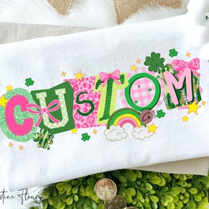 May include: White fabric with the word "CUSTOM" in a colorful, patchwork design. The letters are decorated with pink, green, and yellow accents, including bows, stars, shamrocks, and a rainbow. The design is set against a backdrop of green foliage and gold coins.