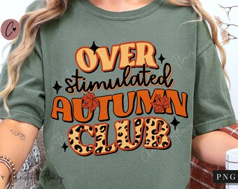 Può includere: T-shirt verde salvia con la scritta "OVER stimulated AUTUMN CLUB" in arancione e nero. La parola "CLUB" presenta un motivo leopardato. La maglietta ha una vestibilità ampia e maniche corte. Il design include foglie autunnali e stelle.