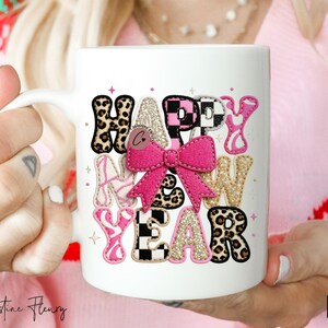 Pode incluir: Caneca de cer&acirc;mica branca com a frase "HAPPY NEW YEAR" num design colorido e divertido. As letras apresentam estampado de leopardo, rosa, glitter dourado e um padr&atilde;o xadrez, com um la&ccedil;o rosa no centro.