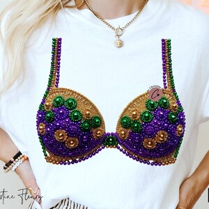 May include: White t-shirt featuring a decorative bra design. The bra is embellished with gold, green, and purple beads. The design is centered on the shirt, with the text "PNG" in the lower right corner.