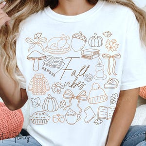 May include: White t-shirt with a brown line art design featuring fall-themed illustrations, including pumpkins, leaves, and a turkey. The words "Fall vibes" are written in a script font.