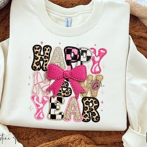 Pode incluir: Camisola creme com as palavras "HAPPY NEW YEAR" num design colorido e divertido. O texto inclui estampados de leopardo, rosa, dourado e padr&otilde;es de xadrez, com um grande la&ccedil;o rosa no centro. Uma pe&ccedil;a festiva e elegante.