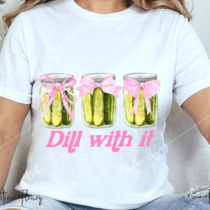 May include: White t-shirt with a graphic of three pickle jars tied with pink ribbons and the text "Dill with it" in pink.