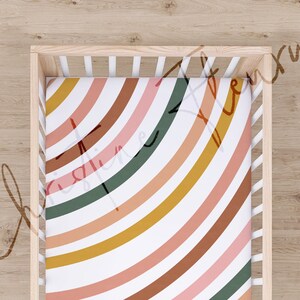 May include: A white cot with a fitted sheet featuring a colourful rainbow pattern. The sheet has a white background with curved stripes in shades of pink, orange, brown, yellow, and green.
