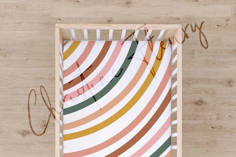 May include: A white cot with a fitted sheet featuring a colourful rainbow pattern. The sheet has a white background with curved stripes in shades of pink, orange, brown, yellow, and green.