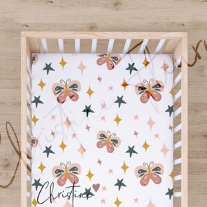 May include: A white fitted cot sheet with a repeating pattern of pink butterflies, gold stars, and dark green stars. The text "Christine" is printed in the bottom left corner.