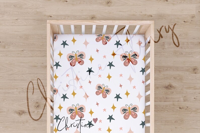 May include: A white fitted cot sheet with a repeating pattern of pink butterflies, gold stars, and dark green stars. The text "Christine" is printed in the bottom left corner.