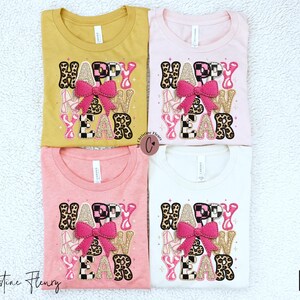 Pode incluir: Quatro camisetas em amarelo mostarda, rosa, coral e branco, cada uma com um design "HAPPY NEW YEAR". O design inclui um la&ccedil;o rosa, estampa de leopardo e detalhes de glitter dourado. As camisetas s&atilde;o dobradas e dispostas em uma superf&iacute;cie branca texturizada.