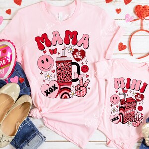 May include: Pink t-shirts with a Valentine's Day theme. The larger shirt says "Mama" with a cartoon juice box, hearts, and flowers. The smaller shirt says "Mini" with a similar design.