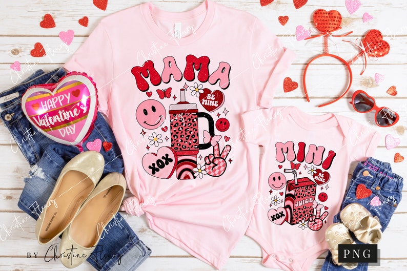 May include: Pink t-shirts with a Valentine's Day theme. The larger shirt says "Mama" with a cartoon juice box, hearts, and flowers. The smaller shirt says "Mini" with a similar design.