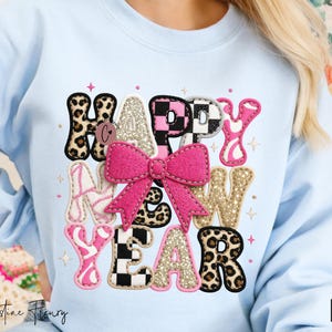 Pode incluir: Camisola azul clara com um design colorido "HAPPY NEW YEAR". O design apresenta um la&ccedil;o rosa, estampa de leopardo e padr&otilde;es xadrez. As letras est&atilde;o em v&aacute;rios estilos e cores, criando um visual festivo.