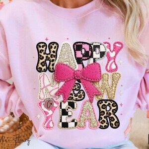 Pode incluir: Um moletom rosa claro com as palavras "HAPPY NEW YEAR" em v&aacute;rias fontes decorativas. As letras apresentam estampa de leopardo, glitter e um la&ccedil;o rosa. O moletom &eacute; uma pe&ccedil;a festiva.