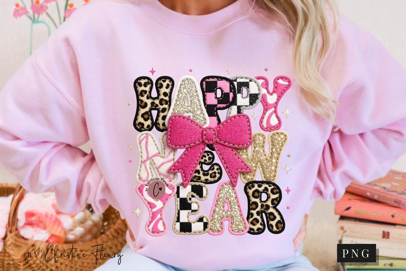 Pode incluir: Um moletom rosa claro com as palavras "HAPPY NEW YEAR" em v&aacute;rias fontes decorativas. As letras apresentam estampa de leopardo, glitter e um la&ccedil;o rosa. O moletom &eacute; uma pe&ccedil;a festiva.