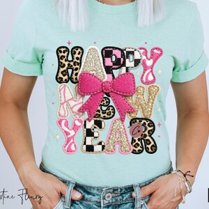 Pode incluir: Uma camiseta verde clara com um design colorido "HAPPY NEW YEAR". O design inclui um la&ccedil;o rosa, letras com estampa de leopardo e detalhes brilhantes. A camiseta &eacute; usada com jeans azuis.
