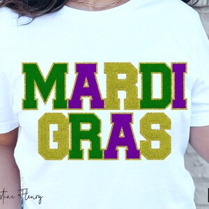 May include: White t-shirt featuring the text "MARDI GRAS" in green, gold, and purple glitter. The design is in a bold, block letter style. The shirt is a crew neck and has a casual style.