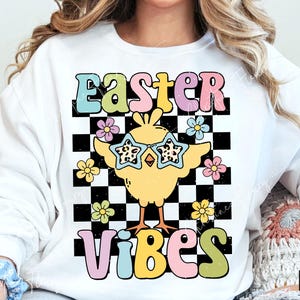 May include: White sweatshirt with a colorful graphic that says "Easter Vibes." The design features a yellow chick wearing star-shaped sunglasses, set against a black and white checkered background, with flowers. A fun, retro-style Easter-themed design.