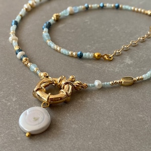 May include: A gold necklace with a large gold clasp and a white pearl pendant. The necklace is made of small beads in shades of blue, white, and gold.