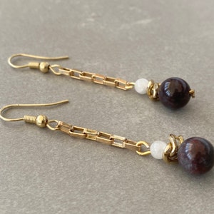 May include: A pair of gold dangle earrings with a white bead and a dark purple bead on each earring.
