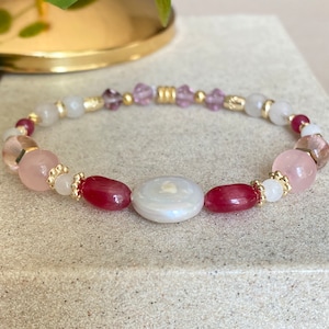 May include: A pink, white, and red beaded bracelet with a large white iridescent oval charm. The bracelet is made with gold-toned beads and spacers.