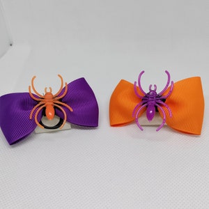 May include: Two Halloween hair bows with purple and orange ribbon and plastic spider accents. The spider on the left bow is orange and the spider on the right bow is purple.