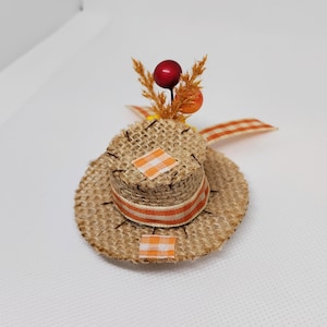 May include: A small, brown burlap hat with orange and white striped ribbon and a small cluster of fall-colored berries on top.