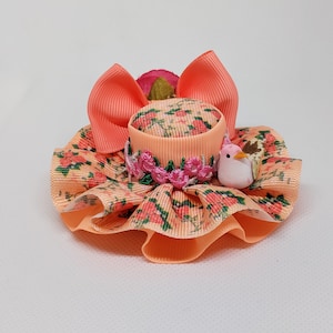 May include: A peach-colored hat with a floral print and a small white bird accent. The hat is made of fabric and has a ruffled edge.