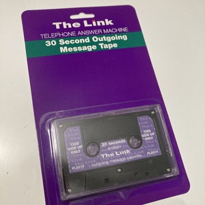 Puede incluir: Una cinta de casete de contestador telefónico "The Link", presentada en un blíster morado. El casete negro tiene texto blanco, incluyendo "30 seconds" y "outgoing message cassette". El embalaje también presenta el texto "30 Second Outgoing Message Tape".